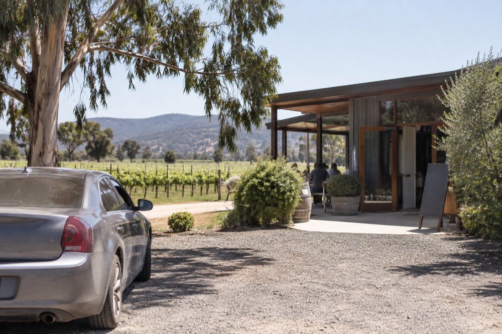 A parked car beside a gravel driveway leading to a cellar door building among vines