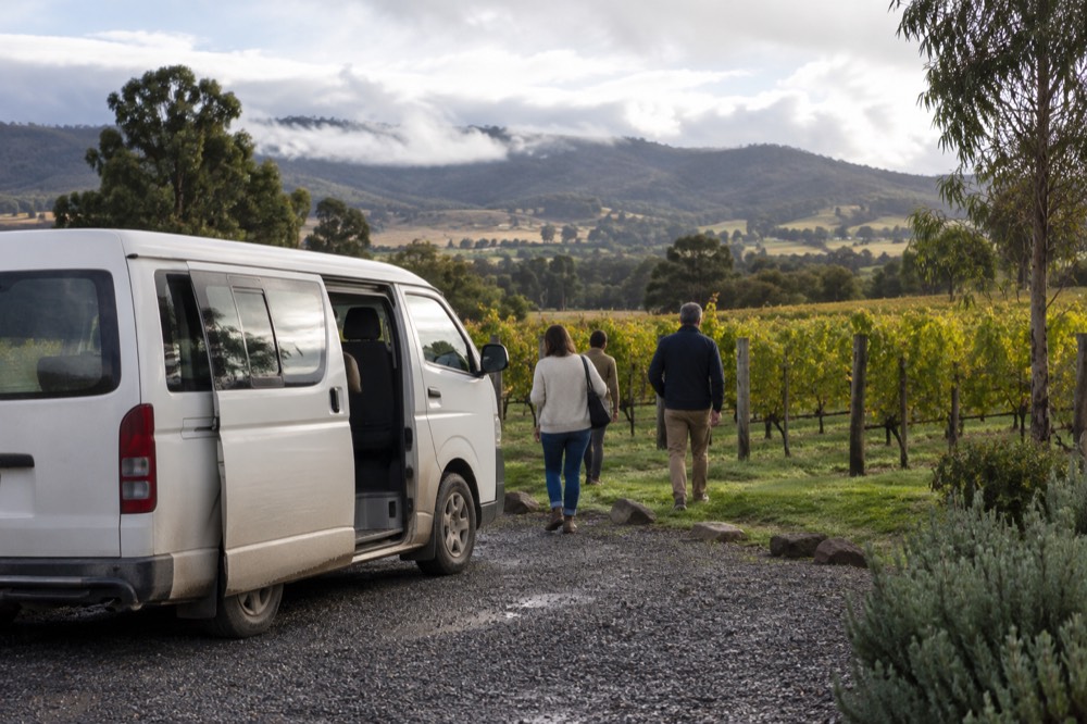 A small tour van parked on a gravel driveway beside a vineyard with rolling green hills
