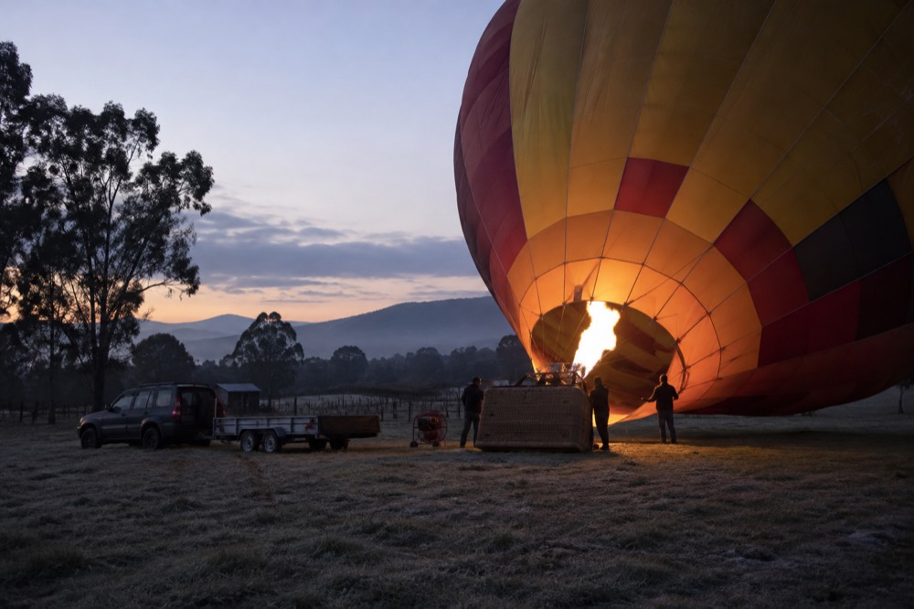 A hot air balloon envelope being inflated on a grassy launch field before dawn, with the crew lit by the burner flame