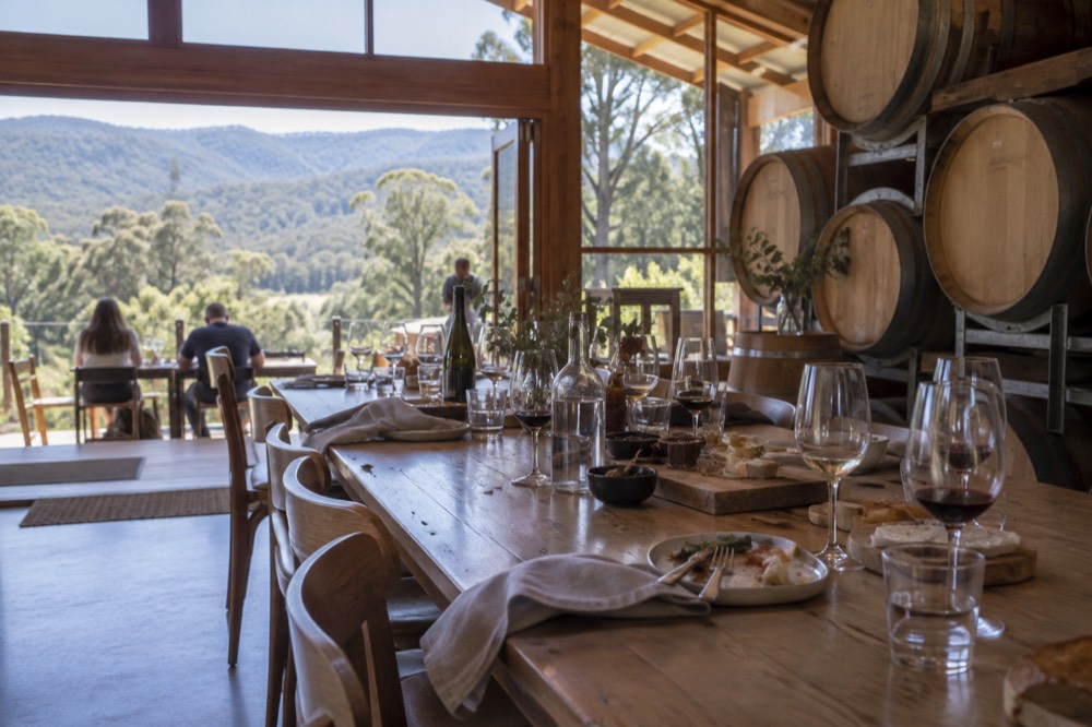 A long table of wine glasses and grazing plates set among barrels inside a bright winery dining room
