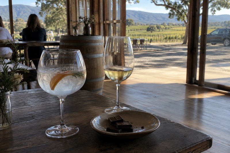 Gin balloon, chocolate squares and a half-drunk chardonnay on a dark timber table in afternoon cellar door light