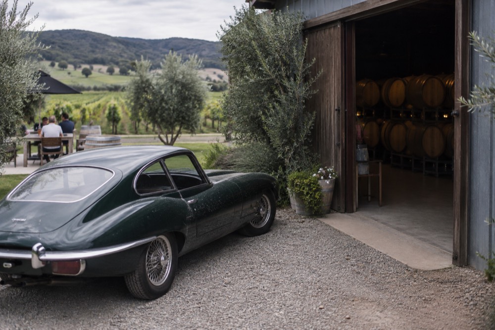 A polished vintage car parked on gravel beside a cellar door entrance with oak barrels visible through the doorway