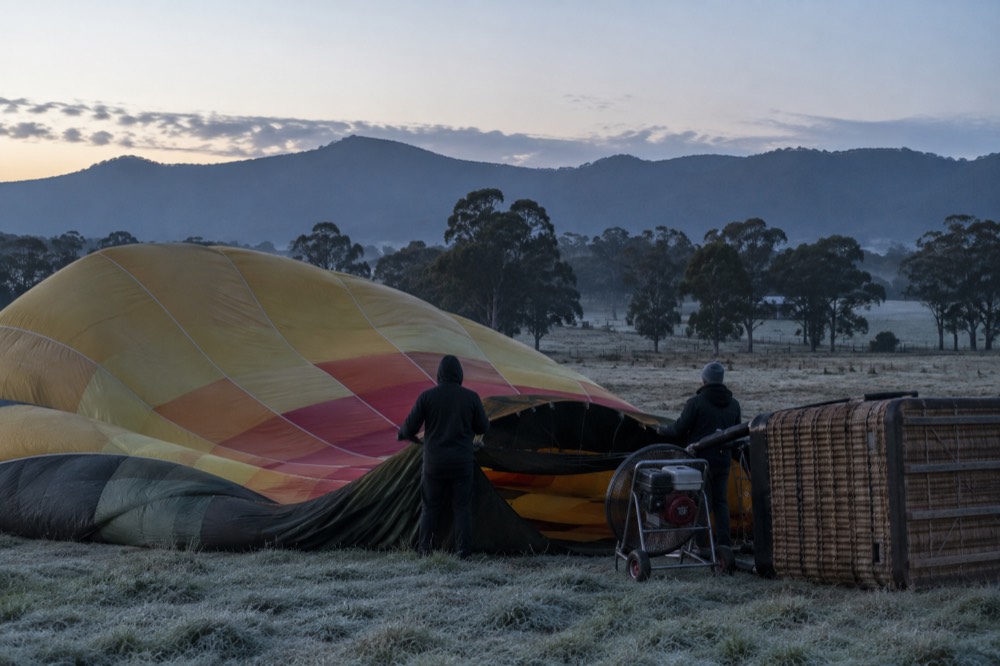 A hot air balloon laid out on a grassy field before sunrise with the envelope being inflated