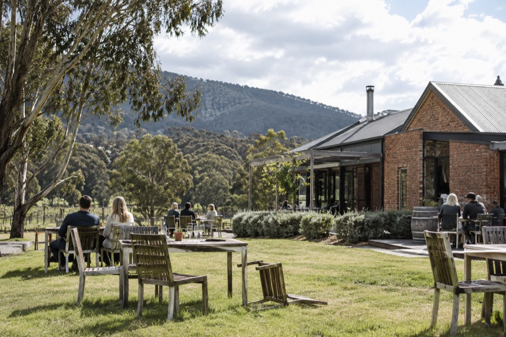 A small winery building with tables on a lawn and hills behind